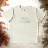 Beige t-shirt with 'Little Windmill Co' text on a wooden surface with decorative elements.