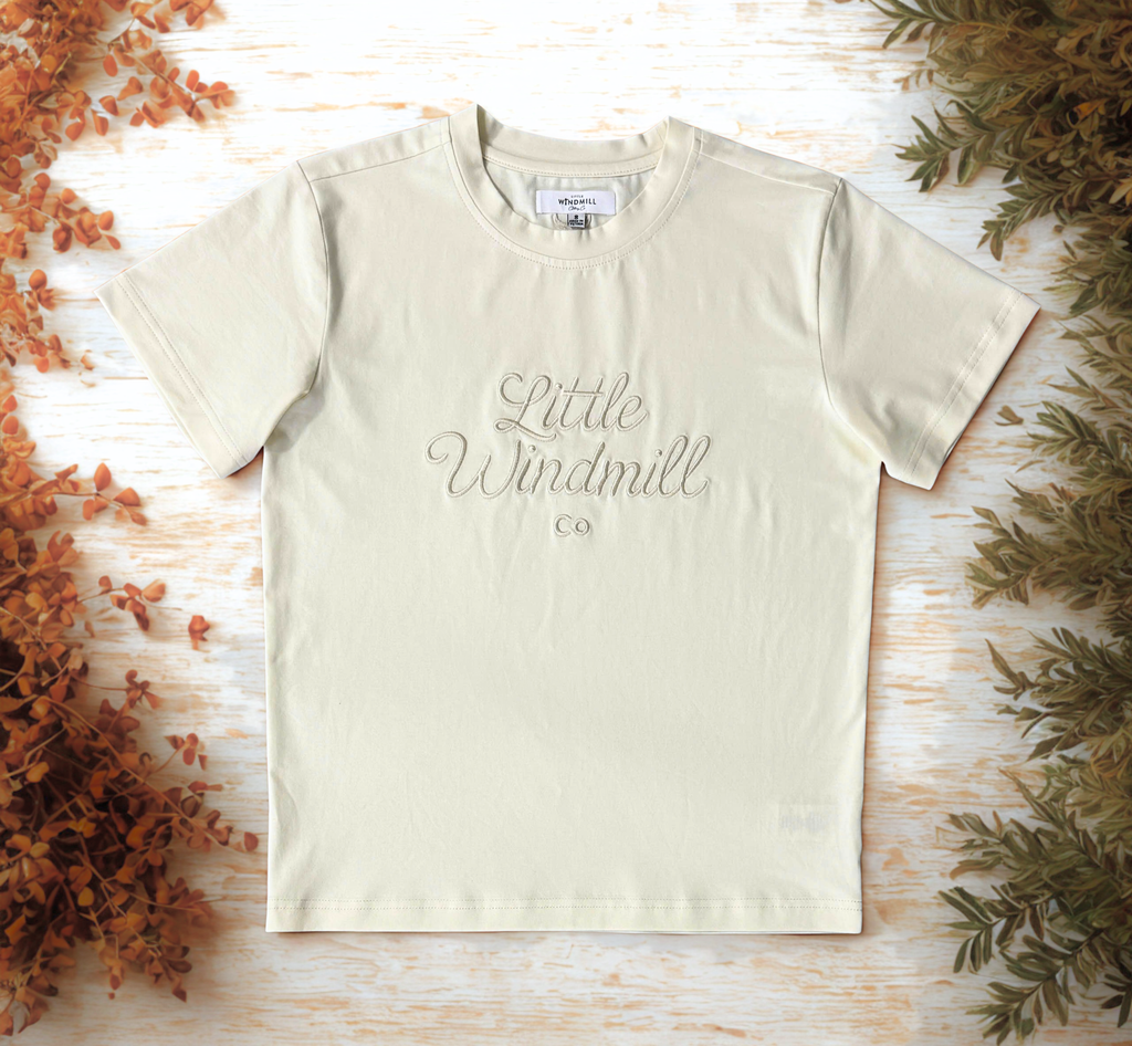 Beige t-shirt with 'Little Windmill Co' text on a wooden surface with decorative elements.