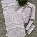 A floral baby travel seat pad set laid out on a concrete floor, featuring a large padded pad, a smaller matching pad, two rolled sections, and a small matching pillow, with a green plant placed on top.