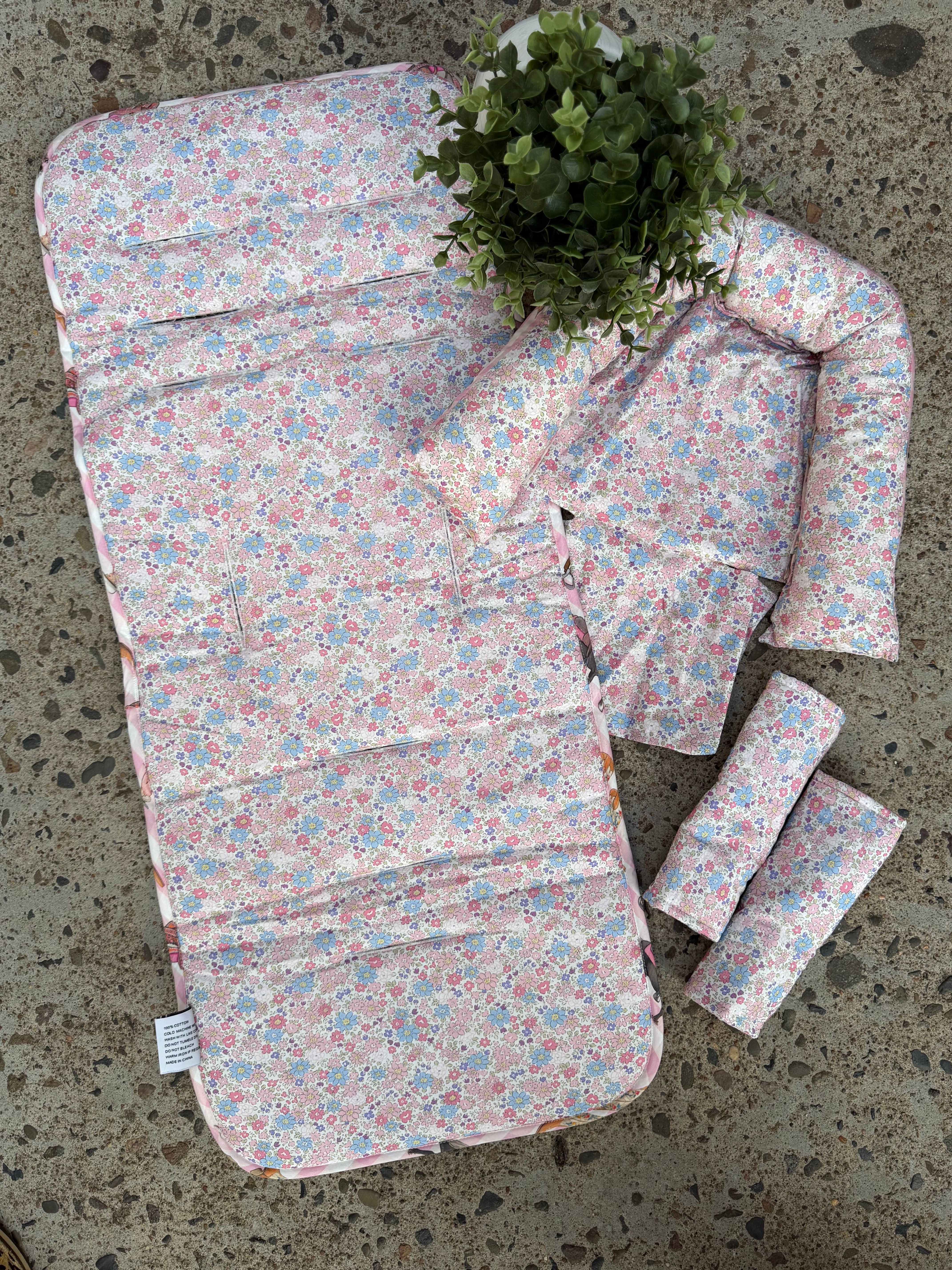 A floral baby travel seat pad set laid out on a concrete floor, featuring a large padded pad, a smaller matching pad, two rolled sections, and a small matching pillow, with a green plant placed on top.