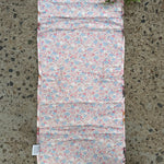 A padded rectangular cushion or pad with a pink, blue, and white floral print, laid flat on a speckled concrete surface with a small green plant visible in the top-left corner.