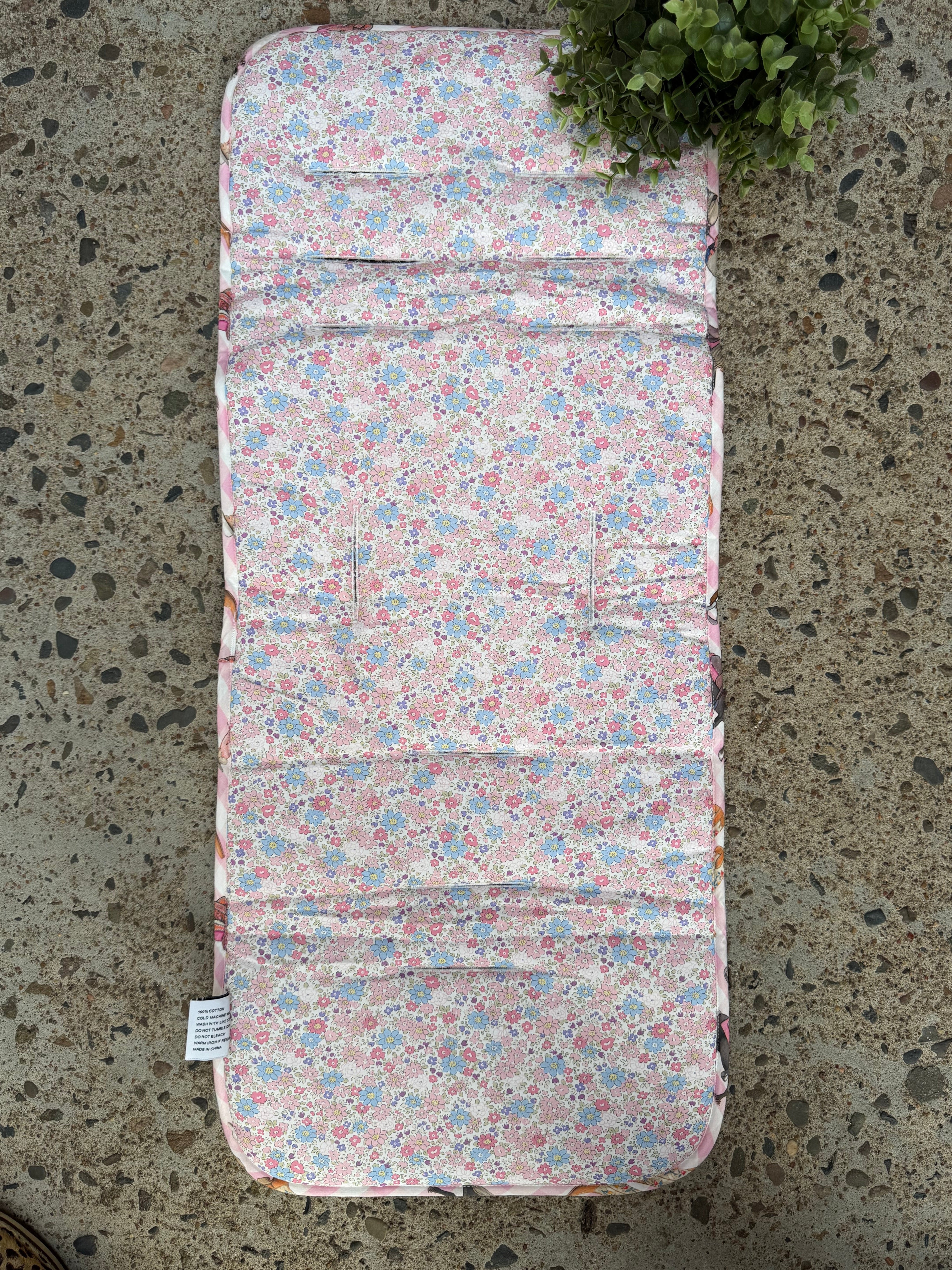 A padded rectangular cushion or pad with a pink, blue, and white floral print, laid flat on a speckled concrete surface with a small green plant visible in the top-left corner.