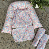 A quilted baby support seat pad with a pink, blue, and white floral print, laid out on a speckled concrete floor next to two matching rolled-up fabric inserts.
