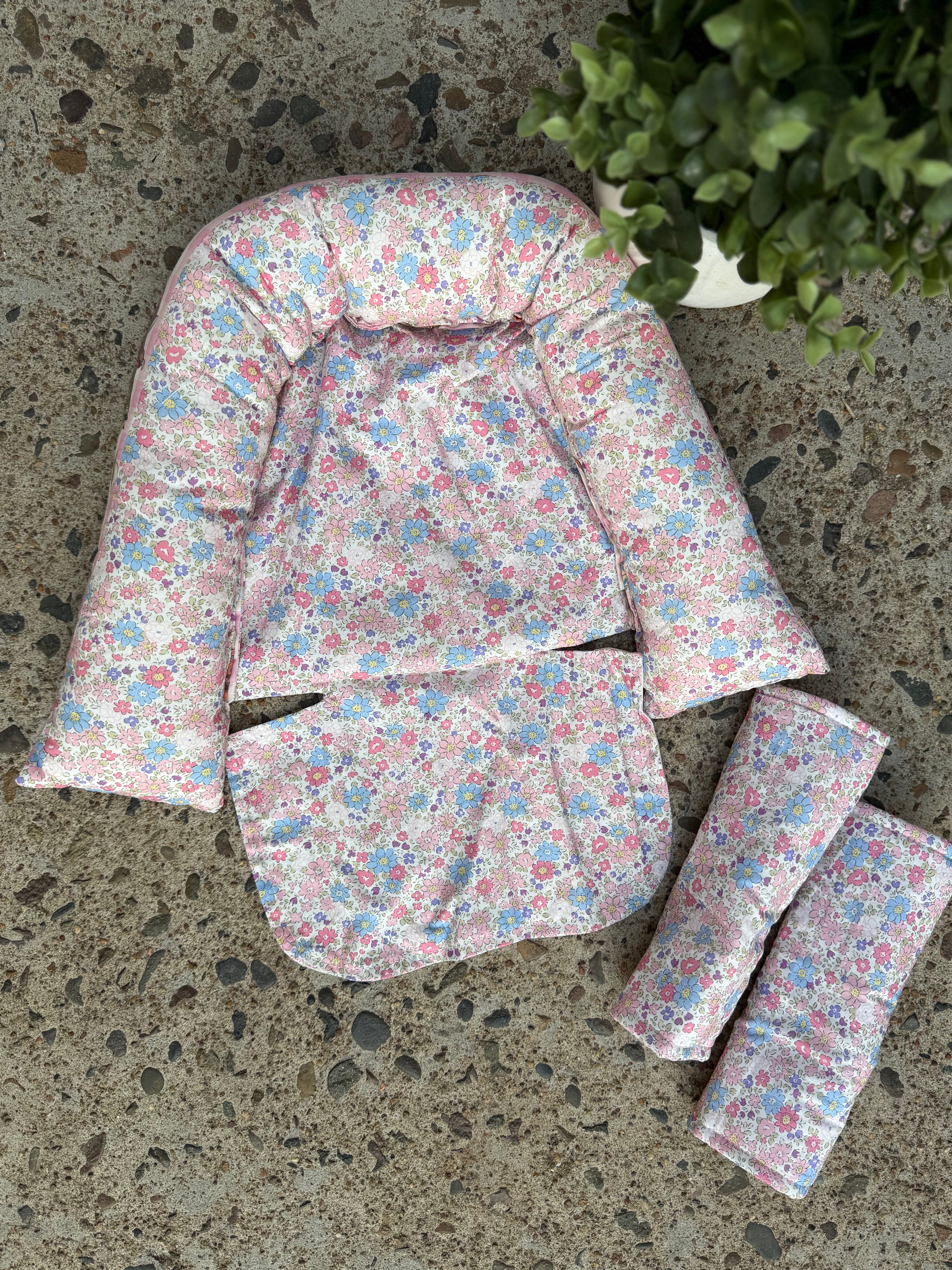 A quilted baby support seat pad with a pink, blue, and white floral print, laid out on a speckled concrete floor next to two matching rolled-up fabric inserts.