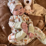 A baby lying on a brown blanket wearing a cream and brown patterned one-piece outfit with matching hat, next to a small plush toy.