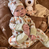 A baby lying on a brown blanket wearing a cream and brown patterned one-piece outfit with matching hat, next to a small plush toy.