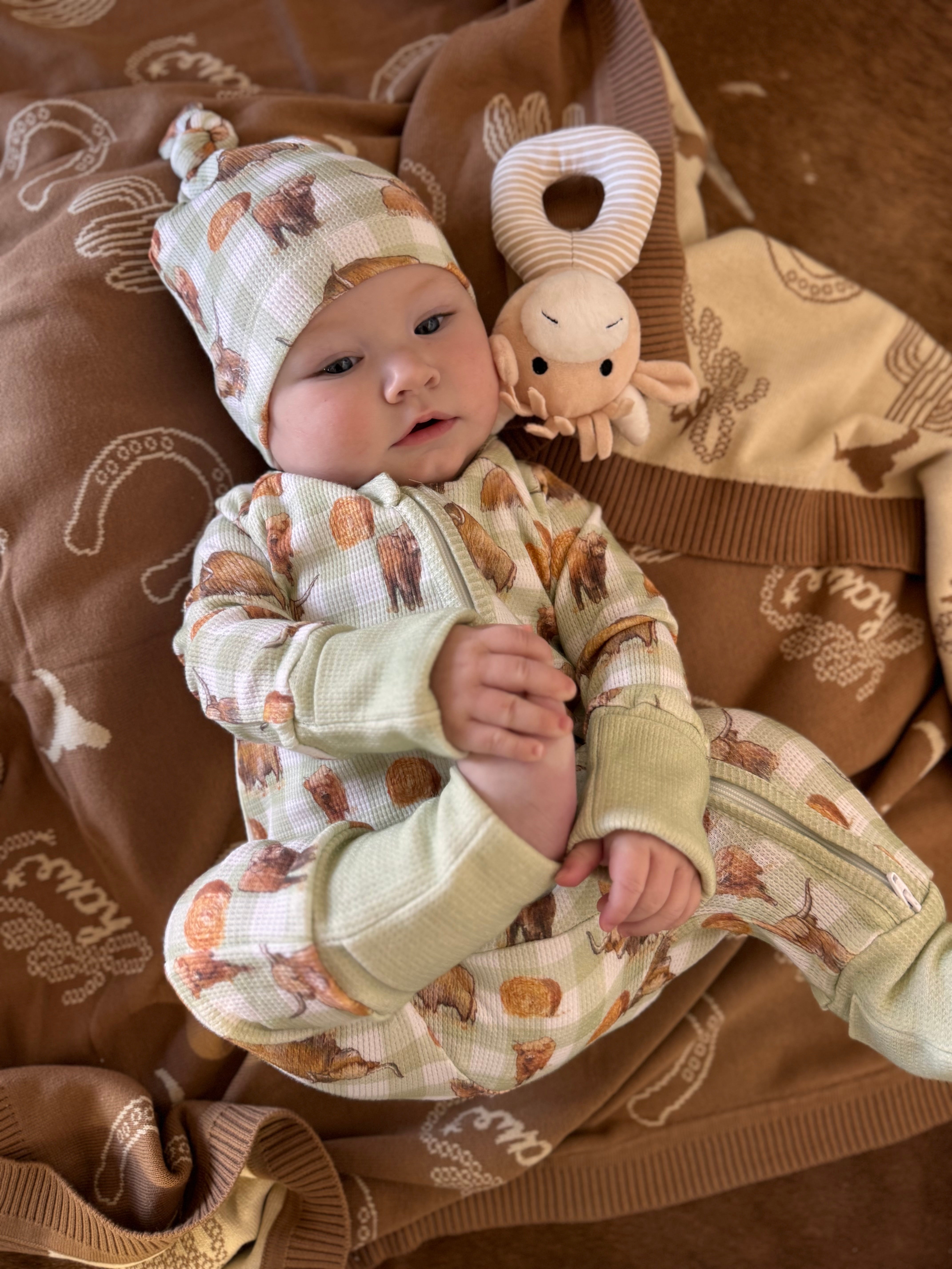 A baby lying on a brown blanket wearing a cream and brown patterned one-piece outfit with matching hat, next to a small plush toy.