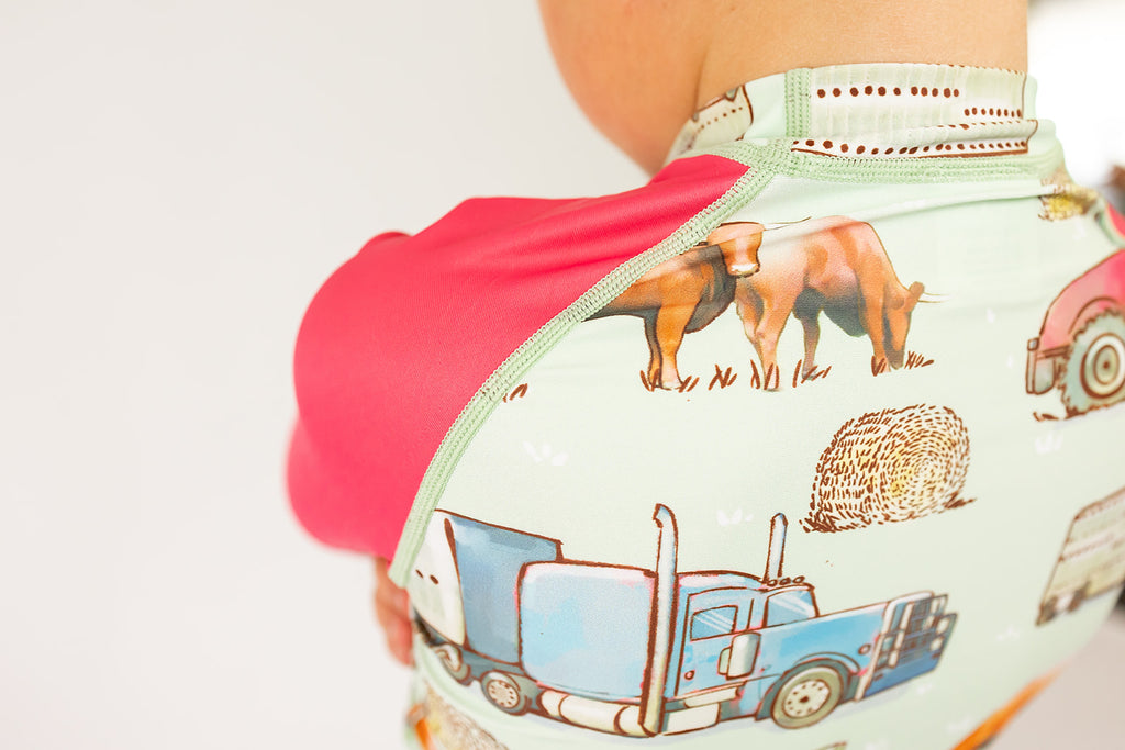 Baby wearing a rash shirt with farm animal and truck design on a plain background